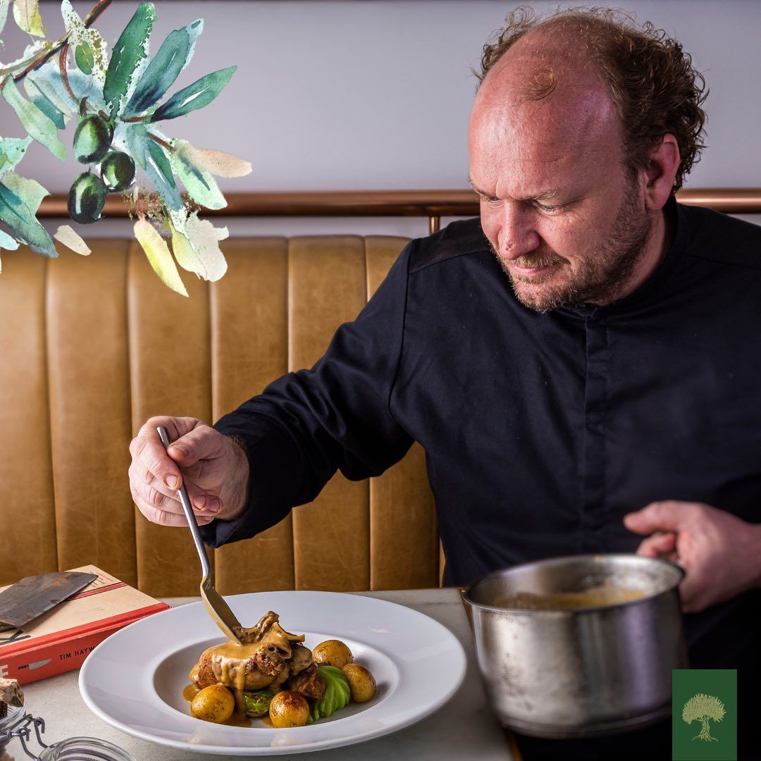 Chef de Cozinha, homem, claro de barba, usando uma doma preta, sentado sobre um estofado segurando uma colher onde despeja sobre um prato de comida um molho, sobre uma mesa de toalha branca com um livro ao lado do prato e na outra mão segura uma panela