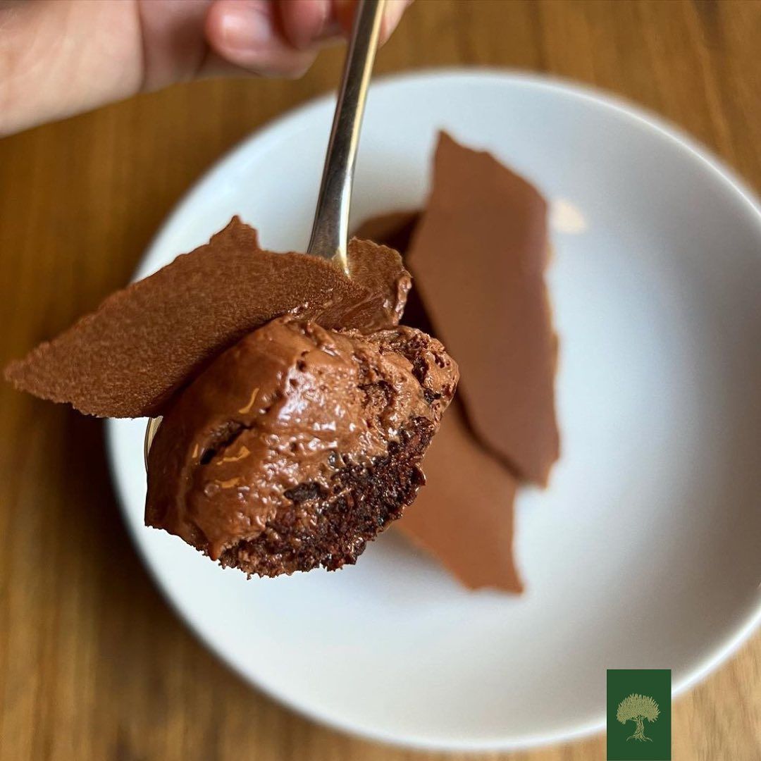 mão segurando uma colher cheia de uma pedaço de uma mousse de chocolate feito com azeite, com uma raspa de chocolate, que está sobre um prato branco em cima de uma mesa de madeira