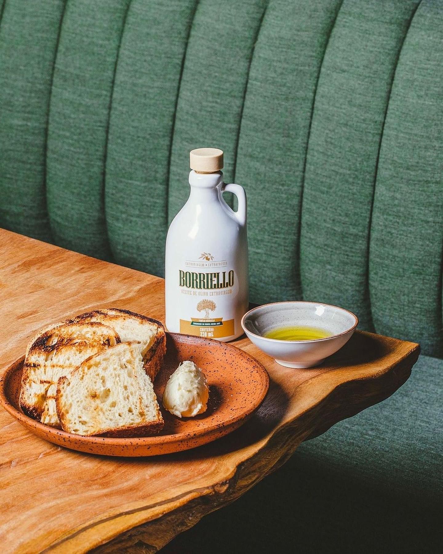 Sobre uma mesa de madeira, uma garrafa branca de azeite borriello ao lado de uma pequena tigela com azeite e de um prato com pães. Ao fundo um estofado verde