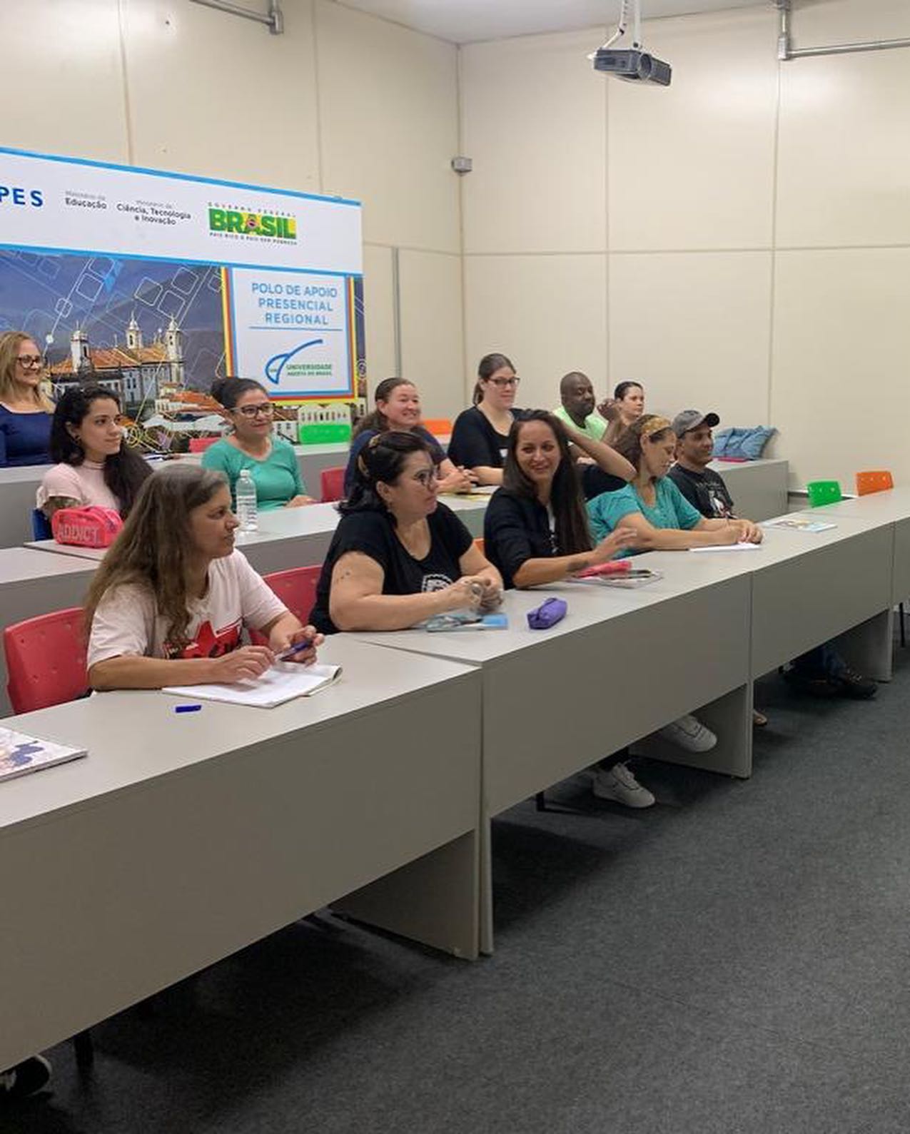 pessoas em uma sala de aula de carpete preto e paredes beges, sentadas e apoiando o seus cadernos e canetas em um balcão cinza e ao fundo um grande cartaz colado a parede